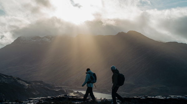 Two hikers walking through a mountainous landscape as sunlight breaks through the clouds