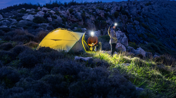 Two campers wearing head torches beside an illuminated yellow tent on a rocky hillside at dusk